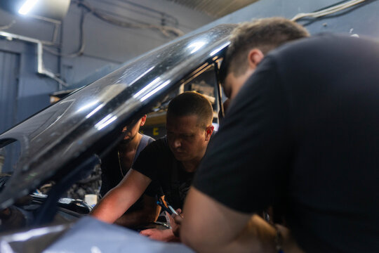 Garage workers gather around faulty automobile with open bonnet. Mechanics carefully observe worn mechanical elements for visible signs of damage