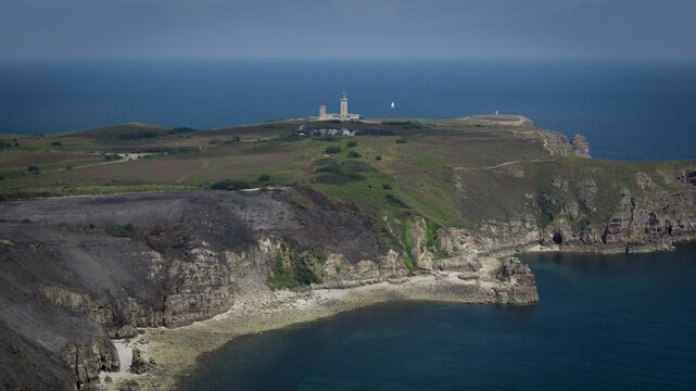 Aerial view of the Cap Frehel lighthouse on the rugged Brittany coastline, showing scorched earth from fire damage alongside green vegetation and the deep blue sea in Plevenon, Brittany, France.