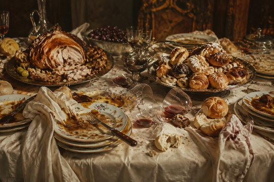 Wide still-life of overloaded banquet table after long meal, torn roast, stacked plates, scattered bread, tipped wine glasses and pastries, gluttony mood