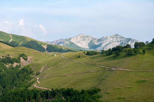 Aerial view of a winding dirt road traversing lush green mountain slopes with rocky peaks in the background in Montenegro.