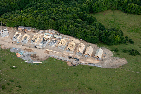 Aerial view of a construction site with several wooden house frames in various stages of development, situated in a rural meadow bordering a dense forest in Montenegro.