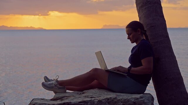 Woman Working on Laptop While Sitting on Coastal Rock Against Palm Tree During Golden Sunset, Peaceful Remote Work by the Sea, Productivity, Freedom, Balance