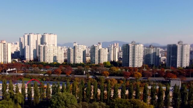 Modern residential skyline borders autumn park, with high-rise apartment towers rising behind colorful trees. Dense urban housing contrasts fall foliage, creating scenic city and nature view.