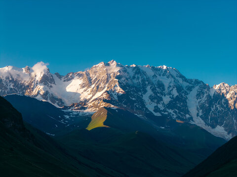 Aerial view of the snow-capped Shkhara mountain range with green slopes under a clear blue sky in Ushguli, Samegrelo-Zemo Svanet, Georgia.