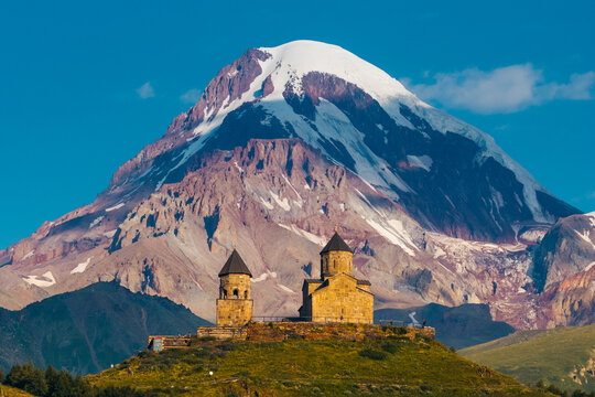Aerial view of Gergeti Trinity Church nestled against the majestic snow-capped Mount Kazbek under a clear blue sky in Oni, Mtskheta-Mtianeti, Georgia.