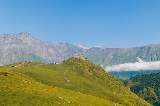 Aerial view of Gergeti Trinity Church perched on a lush green hill surrounded by majestic Caucasus mountains under a clear blue sky in Oni, Mtskheta-Mtianeti, Georgia.