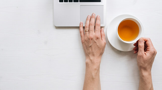 Woman's hands holding tea cup at desk with laptop and trackpad