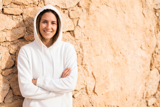 Young woman in white hoodie arms crossed stone wall portrait