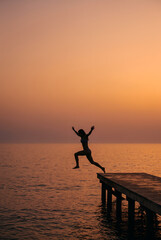 Fototapeta premium Silhouette of woman jumping from wooden pier at colorful sunset