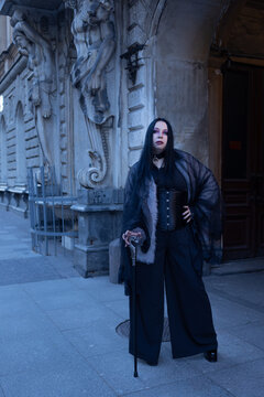Elegant woman in corset, wide black trousers and mesh cape with cane, gothic street style.