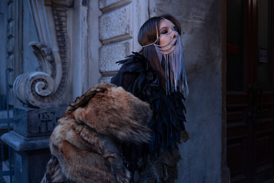 Side view of a woman in a metal chain veil and feathered leather cape, dark party style