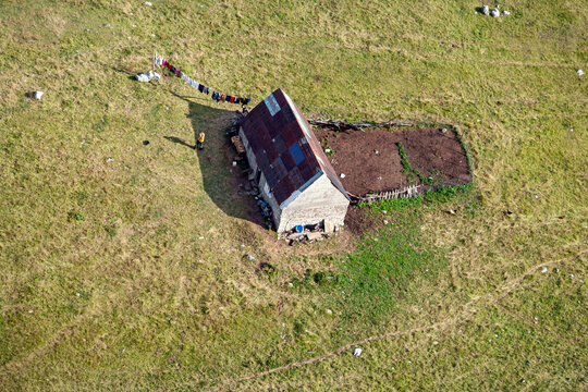 Aerial view of a small rural house with a metal roof, a clothesline with laundry, and a garden plot in a grassy field in Montenegro.