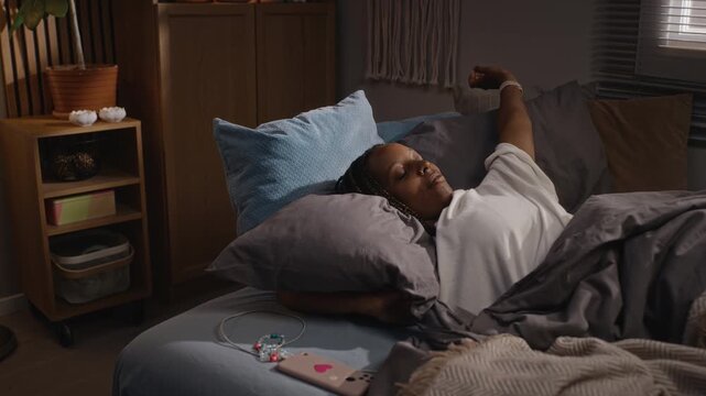 Young adult Black woman waking up in bed, stretching, and immediately checking her smartphone. Perfect for commercial use in campaigns about digital burnout, sleep hygiene, and mental wellbeing