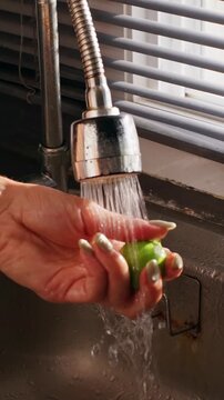 Vertical video. Woman Rinses Individual Green Lime beneath Running Kitchen Faucet in Slow Motion, Fresh Ingredient Cleaning and Healthy Eating