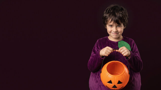 Smiling Boy Wearing a Grape Halloween Costume with Purple Fruit Outfit Holding an Empty Jack-o-Lantern Candy Bucket on a Dark Burgundy Background