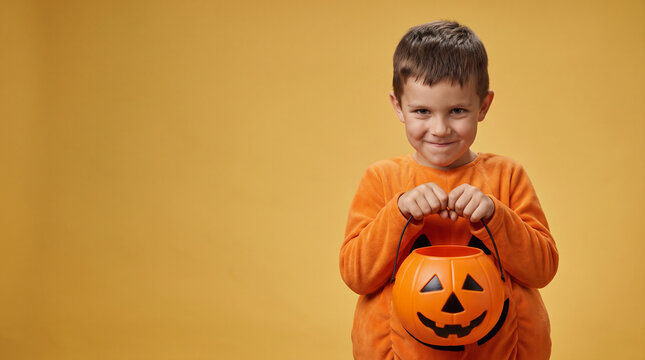 Smiling Boy Wearing an Orange Pumpkin Halloween Costume Holding an Empty Jack-o-Lantern Pumpkin Candy Bucket for Trick-or-Treating on an Orange Studio Background