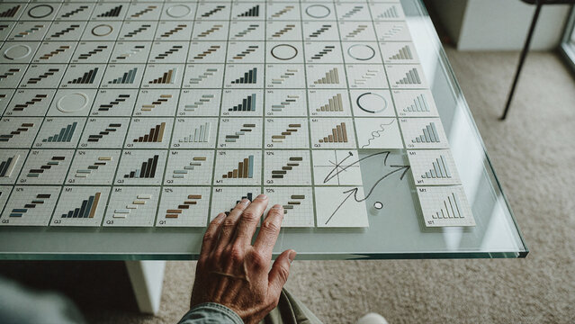 Hand reaches toward organized data charts spread across a glass tabletop in an indoor workspace. Soft natural illumination highlights grid lines, bar graphs, and circular diagrams on white paper.