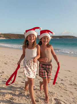 Two Happy Little Girls in Santa Hats Running on Sandy Beach Ocean Shore