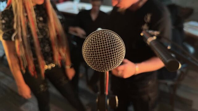 Close up of dynamic microphone on stand with blurred alternative youth with dreadlocks in background preparing for open mic poetry slam or amateur karaoke night in cozy underground indie cafe arc shot