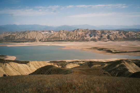 Aerial view of Orto Tokoy Reservoir with arid rolling hills and distant mountain ranges under a clear blue sky in Kyrgyzstan.