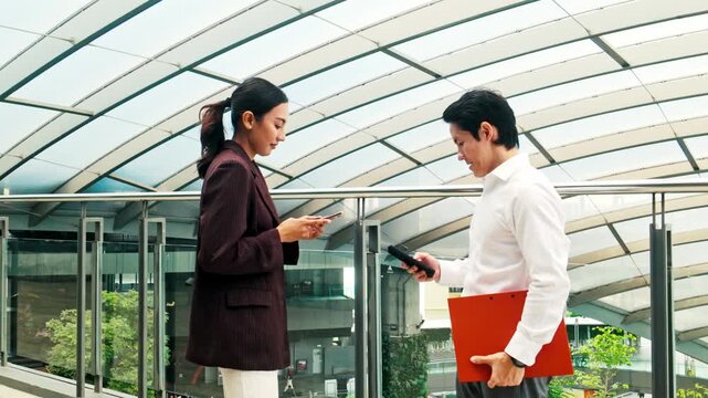 Two professionals under a modern glass canopy compare smartphones during a work meeting, one holding an orange folder on an elevated walkway in a contemporary office setting.