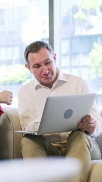 Man sitting on a couch with a laptop in a bright modern office, engaged in conversation near large windows