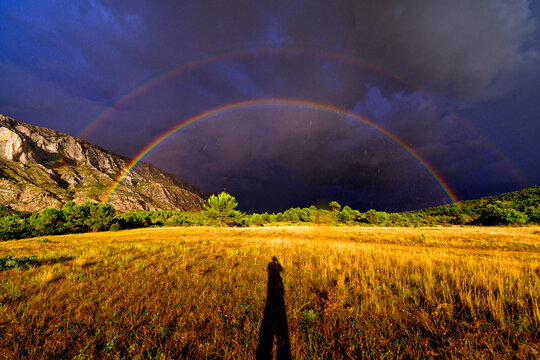 Person casting shadow under double rainbow in mountains