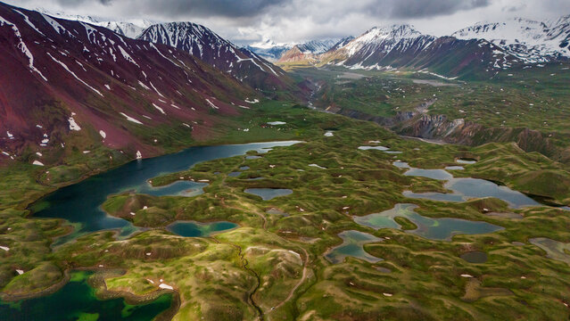 Aerial view of Tulpar Lake with its unique turquoise ponds, rolling green hills, and snow-capped mountains under a dramatic cloudy sky in Alay District, Osh Region, Kyrgyzstan.