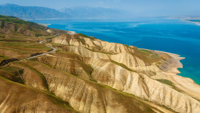 Aerial view of Orto Tokoy Reservoir with its winding road along arid, undulating hills and vibrant blue water under a clear sky in Kyrgyzstan.