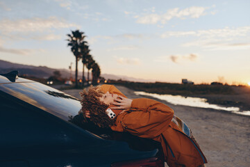 Naklejka premium Woman lifestyle at sunset beachside, curly hair and orange jacket leaning on a parked car hood, relaxed hand on hair, warm golden light, travel vacation mood with blurred ocean waves in soft