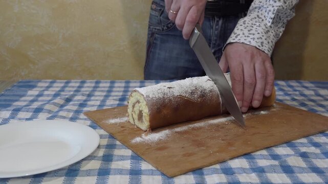Man cutting a sweet homemade biscuit roll cake with a knife, Slicing a delicious sweet pastry roll on a checkered tablecloth in a home kitchen