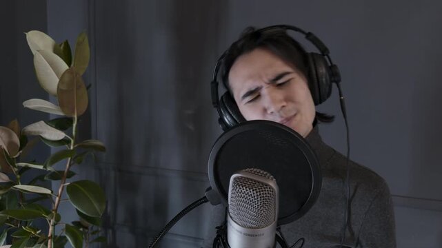 Focused young Asian male voice actor wearing headphones reading intense dramatic monologue into a condenser microphone with pop filter inside modern home recording studio close up handheld shot