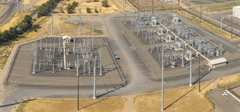 Aerial view of an electrical power substation with transformers, metal structures, and power lines within a fenced gravel compound in the dry landscape of Hermiston, Oregon, United States.