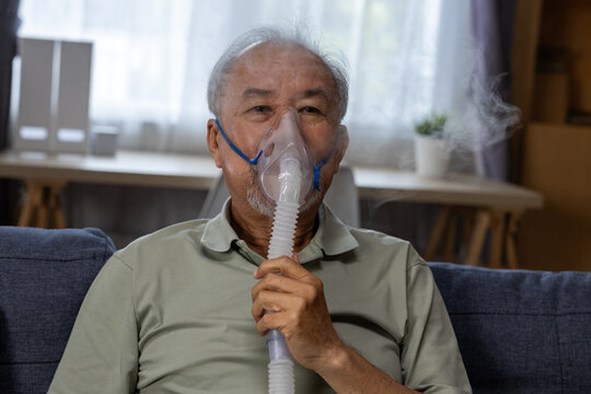Elderly Asian man using nebulizer mask at home, receiving respiratory treatment while seated on sofa with calm expression and visible vapor in domestic setting