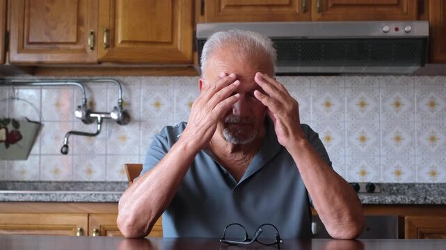 Exhausted elderly man taking off his glasses and rubbing his temples while sitting at a kitchen table, experiencing a painful migraine, stress, and eye strain from overworking