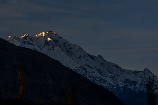 View of the snow-capped peak of Rakaposhi illuminated by golden sunset light above dark mountain slopes in Pakistan.