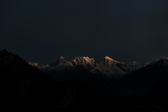 View of Spantik peak with snow-capped summits illuminated by the first light of dawn against a dark mountain range in Nagar, Pakistan.