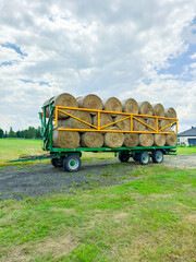 Farm tractor with a hay trailer filled with round bales parked on a green field under a cloudy sky in summer © DSMT