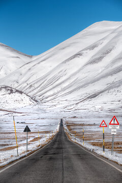 View of the road leading towards the snow-covered mountains of Castelluccio under a clear blue sky in Castelluccio, Umbria, Italy.