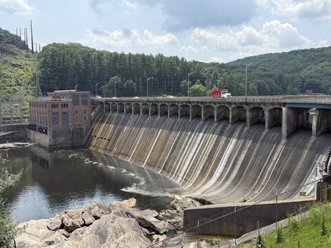 View of Stevenson Generating Station hydroelectric dam with water flowing over the concrete spillway and a roadway crossing the top in Monroe, Connecticut, United States.