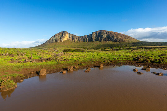 View of a tranquil pond reflecting the blue sky in a lush green valley with a prominent rugged mountain range in the background, Easter Island, Valparaiso, Chile.