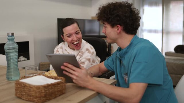 Male caregiver sharing a funny video on a digital tablet with a happy woman in a wheelchair, fostering a positive and supportive relationship in a home care setting