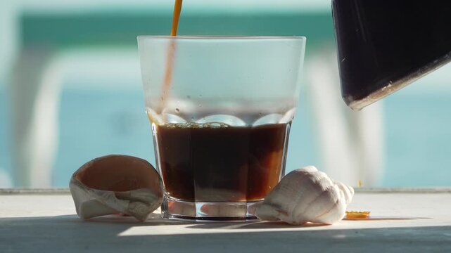 Black coffee fills a transparent tumbler positioned between two oceanic seashells on a bright seaside table. Static medium shot.