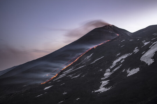 View of Mount Etna erupting with glowing lava flows descending the dark volcanic slopes, patches of snow, and smoke plumes in Catania, Sicily, Italy.