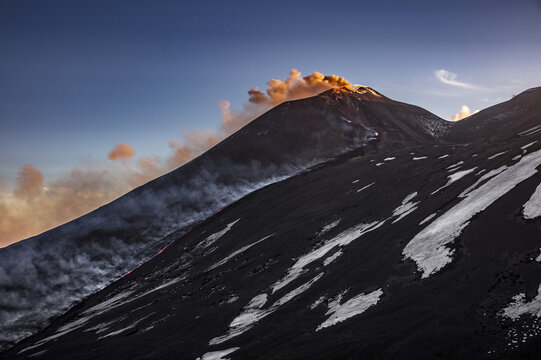 View of Mount Etna erupting with volcanic smoke and ash against a clear sky, with snow patches on the dark volcanic slopes during golden hour in Catania, Sicily, Italy.