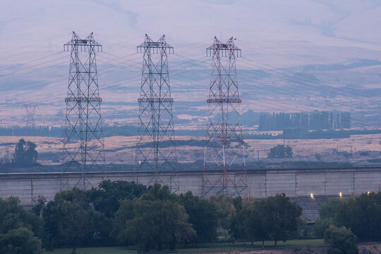 View of The Dalles Dam with large metal transmission towers standing in front of rolling hills and a concrete wall under a soft sky in The Dalles, Oregon, United States.