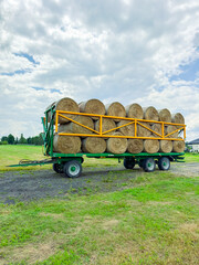 A green trailer loaded with round hay bales is parked on a field under a cloudy sky at a rural farm © DSMT