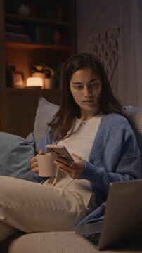 Vertical video of young adult Caucasian woman sitting on sofa at night, holding mug and scrolling on smartphone near laptop. Footage representing digital burnout or remote work fatigue concepts