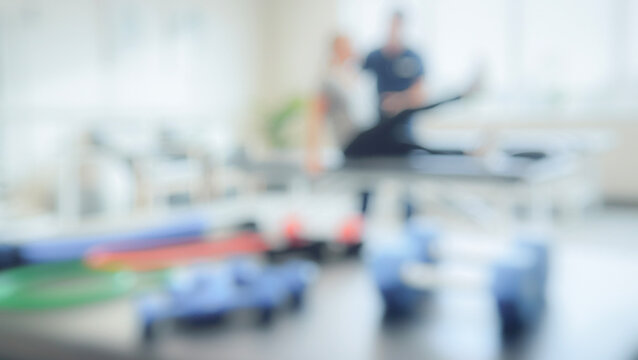 A blurred image of a physical therapist assisting a patient with rehabilitation exercises in a modern clinic setting filled with exercise equipment.