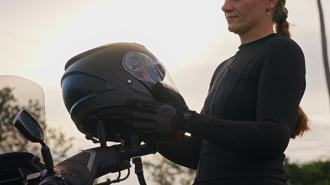 Female Motorcyclist Places Black Full Face Helmet on Motorcycle, Outdoors at Sunset, Showing Rider Preparation and Road Safety